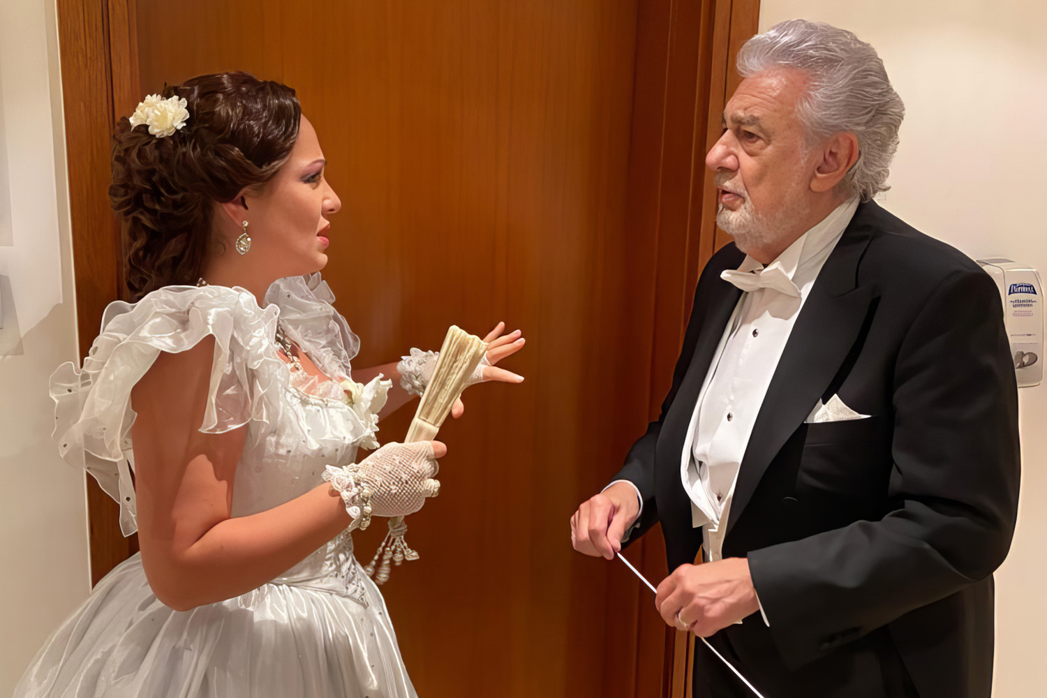 Performer in a white bridal-style gown with a fan beside a conductor in a tuxedo